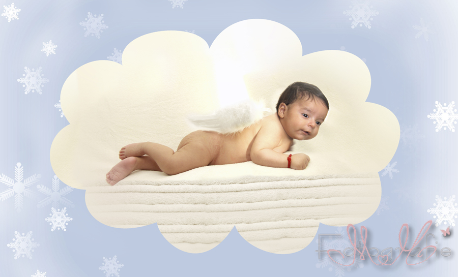 Auf einem Stapel weißer Handtücher liegt das Baby auf dem Bauch mit Engelsflügelchen, umgeben von einer hellblauen Wolke mit weißen Sternen.