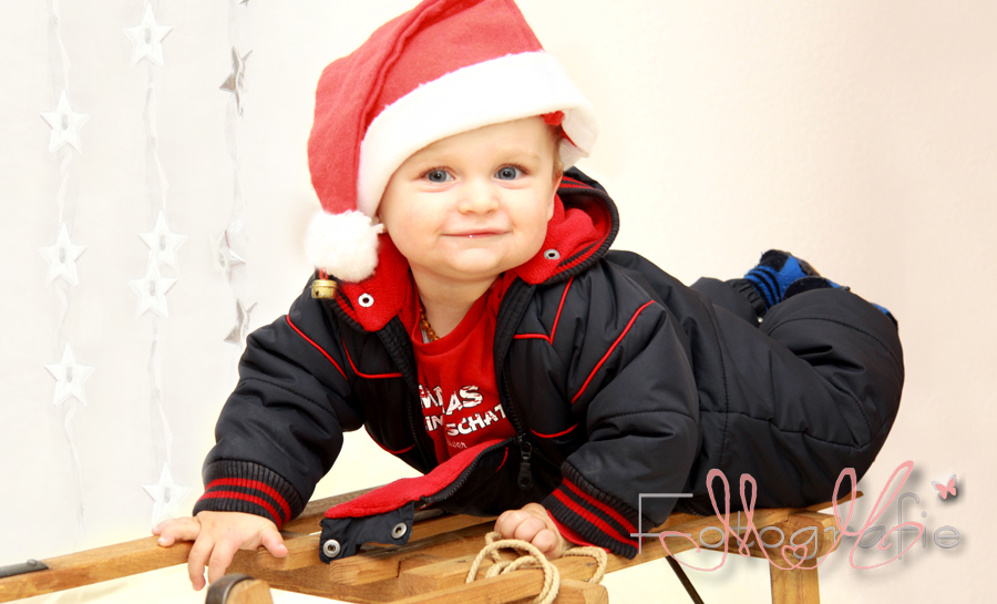 Babyfoto eines kleinen Jungen, er liegt auf einem Schlitten, trägt einen blauen Skianzug und hat eine lustige Weihnachtsmannmütze auf dem Kopf.
