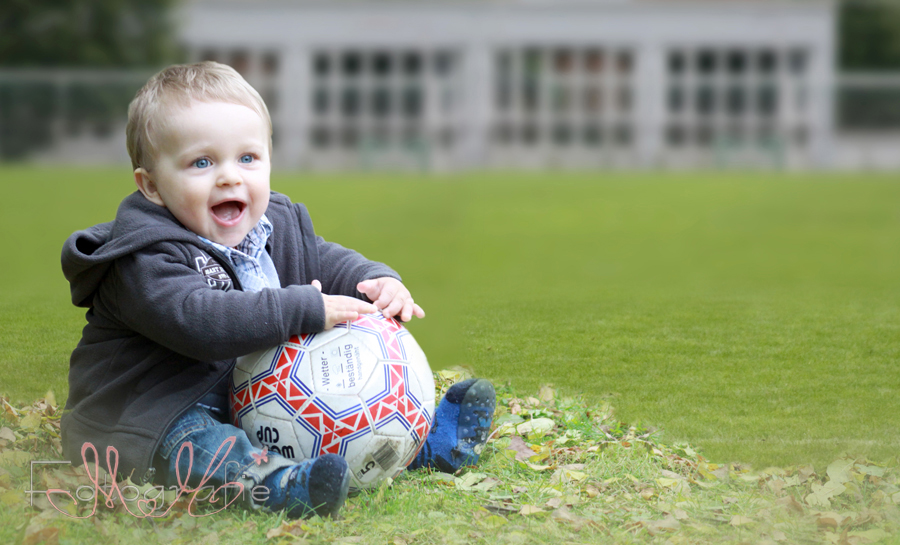 Farbfotografie eines Kleinkindes, es sitzt auf dem Rasen eines Fußballfeldes und hält einen weiß-roten Fußball fest. 