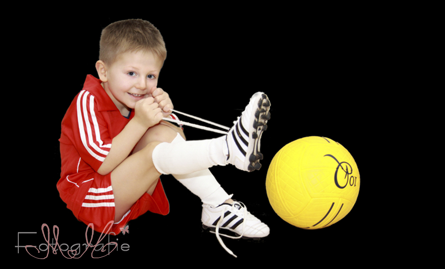 Fotografie eines Kleinkindes, der kleine Junge zieht seine Fußballschuhe an, er trägt ein rotes Fußballtrikot und hat einen gelben Fußball neben sich liegen.