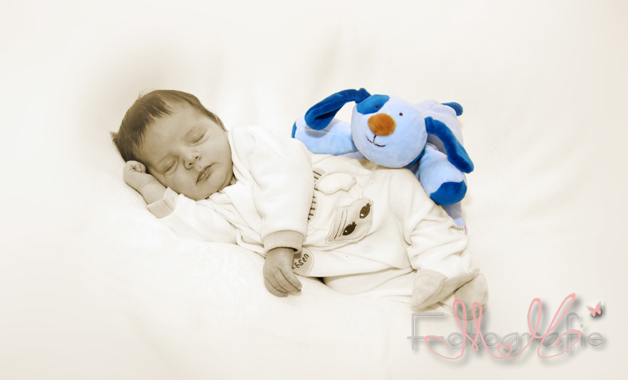 Sepia-Fotografie eines Neugeborenen, es liegt schlafend auf der Seite, hinter ihm schaut ein blaues Kuscheltier hervor.