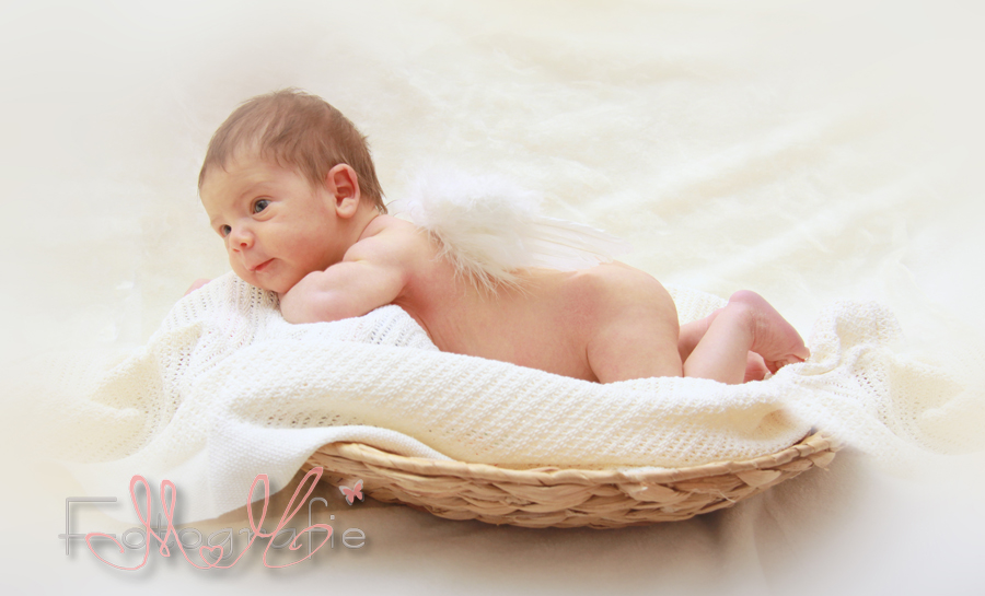 Bild eines Neugeborenen, das nackig auf dem Bauch in einem runden Körbchen liegt, auf einer weißen Strickdecke. 