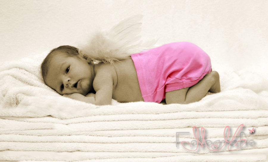 Ein neugeborenes kleines Mädchen liegt auf einem Stapel weißer Handtücher, auf dem Rücken sind kleine Engelsflügel, es trägt eine pinkfarbene Hose.